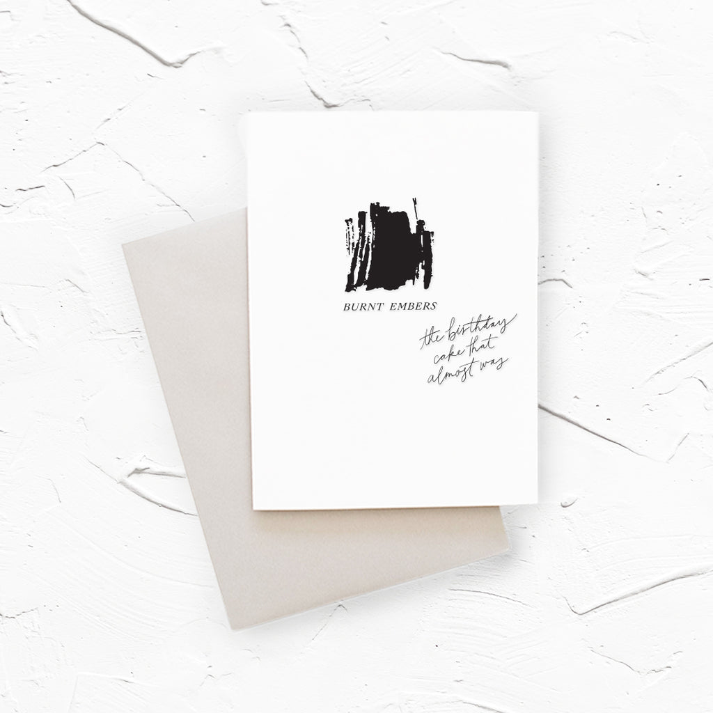 White card with a black square in center with black text saying, “Burnt Embers The Birthday Cake That Almost Was”. A gray envelope is included.