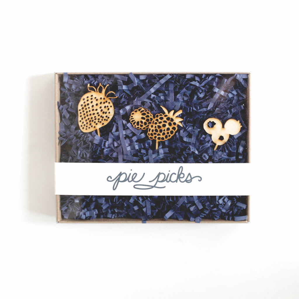 Gold metal pie picks in the shapes of strawberries, raspberries and blueberries. Clear packaging with blue stuffing and white label with blue text saying, “Pie Picks”.