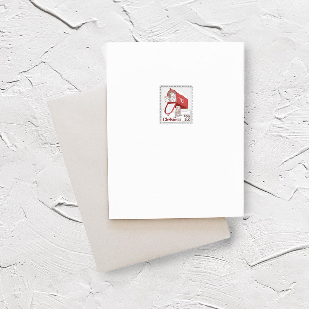 White card with design of a postage stamp with the image of a red mailbox with many envelopes comig out of it. Red text saying, “Christmas”. A gray envelope is included.