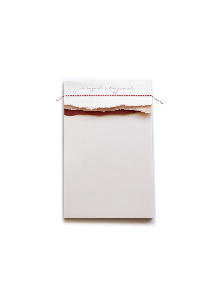 Ivory notepad with pink and red torn edging across the top with red text saying, “The Magic is in Doing the Work”.
