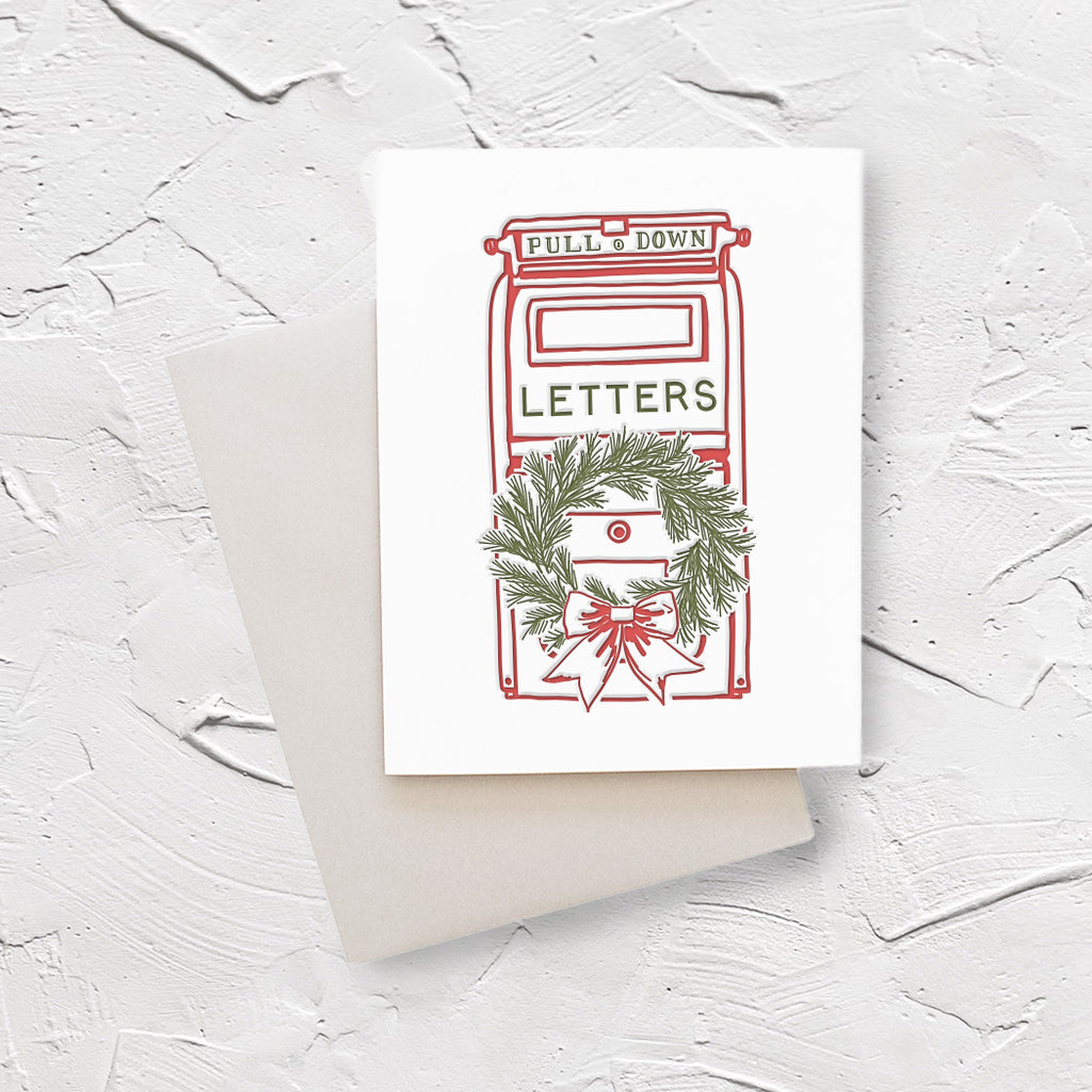 White card with image of vintage red mailbox with holiday green wreath on front with red bow. Writing on mailbox saying,