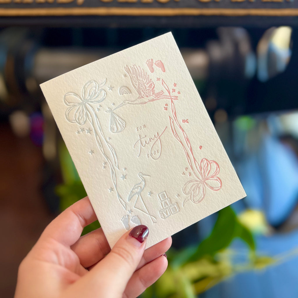 White card with blue and pink embossed images of baby things: stork, blocks, baby feet, stars and rainbows. Text saying,